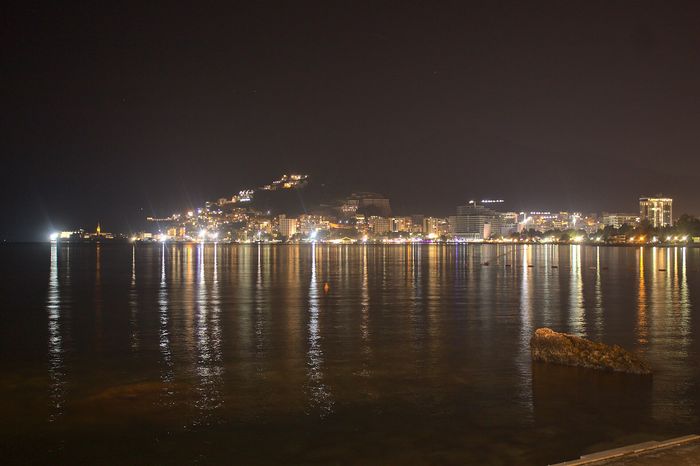 Noćna obala Budve, sa svetlima obale odraženim u vodi.